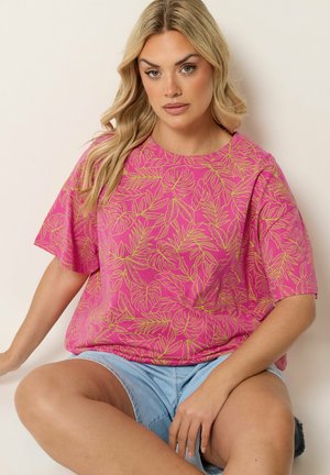 Blonde vrouw zittend, gekleed in een roze shirt met gele bladpatronen en lichtblauwe spijkershorts tegen een effen achtergrond.