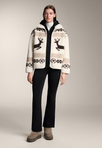 Kurtka polarowa w kremowo-brązowym kolorze, ozdobiona wzorami reniferów i geometrycznymi akcentami, zestawiona z czarnymi dopasowanymi spodniami i beżowym obuwiem.