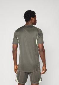 Camiseta deportiva en verde olivo oscuro, de mangas cortas, con acentos en verde claro en los hombros. Tejido suave y ligero con un dobladillo curvado.