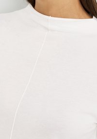 Vit höghalsad topp gjord av mjuk bomull, med en sömdetalj nedanför framsidan och en slät, odekorerad textur.