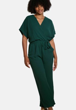 Frau in einem dunkelgrünen, kurzärmligen Jumpsuit mit V-Ausschnitt und gebundenem Bund, stehend vor einem schlichten weißen Hintergrund.