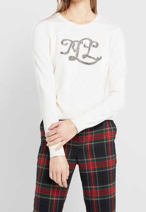 Kvinde iført en hvid langærmet sweater med dekorative "RL" initialer, kombineret med røde og grønne ternede bukser, stående foran en ensfarvet baggrund.