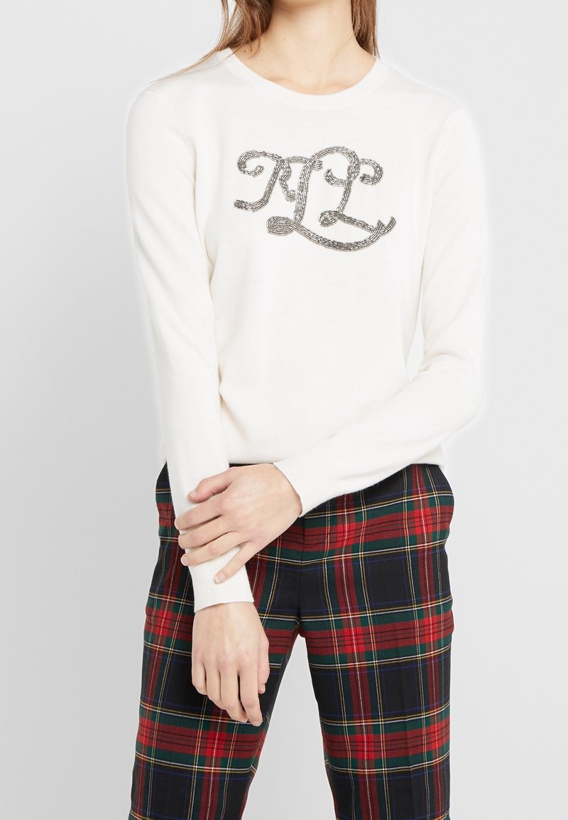 Femme portant un pull blanc à manches longues avec des initiales décoratives « RL », assorti à un pantalon à carreaux rouge et vert, debout devant un fond uni.