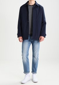 Marineblå letvægtsjakke med knaplukning, båret over en stribet skjorte, kombineret med lyseblå jeans og hvide sneakers. Minimalistisk design.