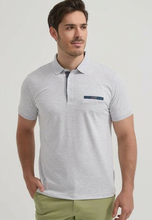 Homme portant un polo à manches courtes gris clair avec un passepoil foncé sur la patte de boutonnage et la poche, associé à un pantalon vert clair, regardant vers la droite.