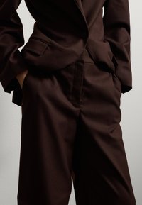 Blazer marrón a medida con pantalones a juego. La tela es suave con un ligero brillo, cuenta con un solo botón y bolsillos laterales.