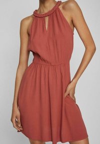 Ein rustfarbenes Halterneck-Kleid mit geflochtenem Ausschnitt, schlanker Taille und fließendem Rock. Der Stoff hat eine strukturierte, zerknitterte Optik.