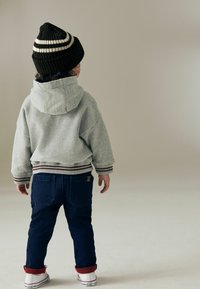 Grau-gestreifter Kapuzenpullover mit gerippten Bündchen, kombiniert mit dunkelblauen Jeans und weißen Sneakers mit roten Akzenten, von hinten betrachtet.