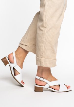 Feet wearing white low-heeled sandals with ankle straps, paired with beige rolled-up corduroy pants, shown in side view.