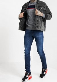 Mężczyzna ubrany w czarną dżinsową kurtkę, szary sweter z ciemnym i czerwonym paseczkiem, wąskie niebieskie dżinsy oraz czarne trampki z czerwonymi i białymi akcentami.