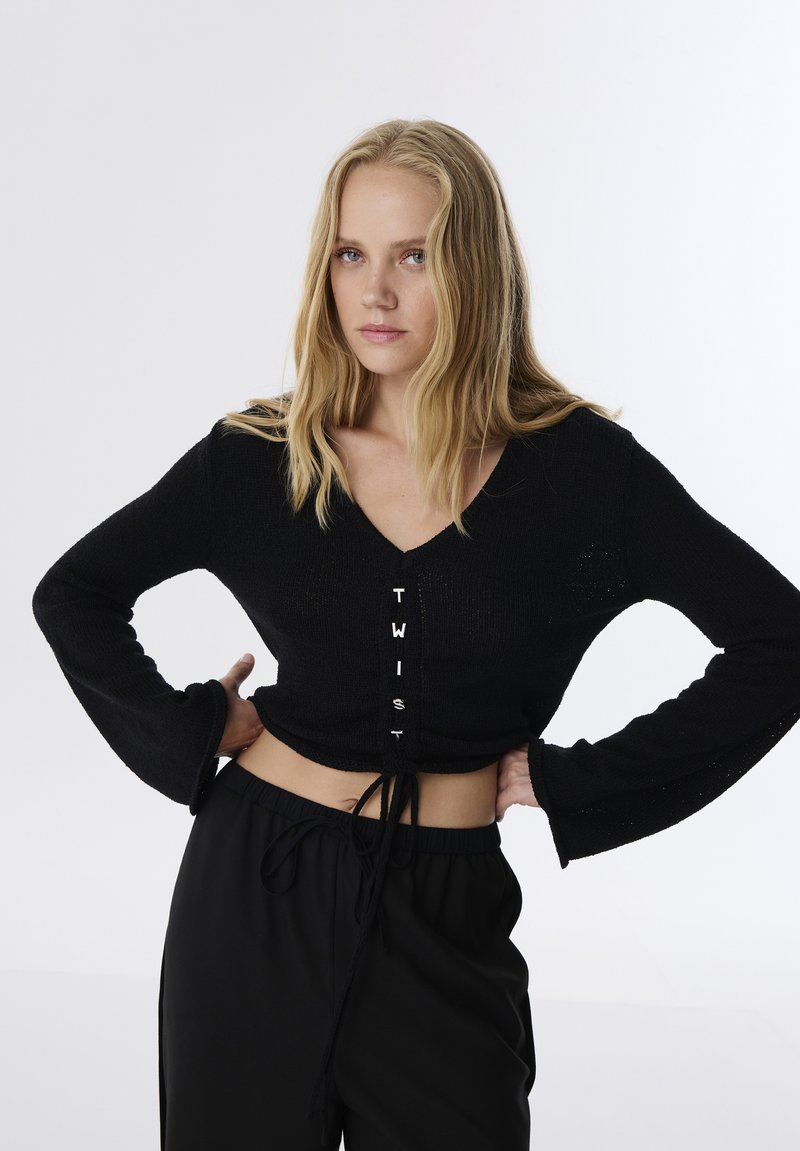 Zwarte cropped sweater met een V-hals, lange trompetmouwen en knopen aan de voorkant met de tekst "TWIST." Gecombineerd met zwarte broek met trekkoord.