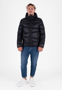 Doudoune noire avec zip à l'avant, capuche et design matelassé. Portée par-dessus un jean bleu et des baskets blanches à lacets. Fond uni.