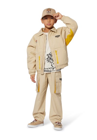 Veste beige avec des accents jaunes, accompagnée d'un pantalon assorti. T-shirt blanc avec un graphique de lion noir. Casquette beige et chaussures avec un détail de tigre.