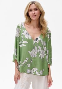 Zielona bluzka w kwiaty, wykonana z lekkiego materiału, z dekoltem w kształcie litery V, rękawami 3/4 oraz luźnym krojem, zdobiona czarno-białymi wzorami kwiatowymi.