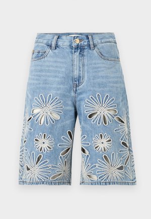 Kobieta stoi z ręką w kieszeni, ubrana w biały top na ramiączkach i dżinsowe szorty do kolan z dużymi wyhaftowanymi wycięciami w kształcie kwiatów.