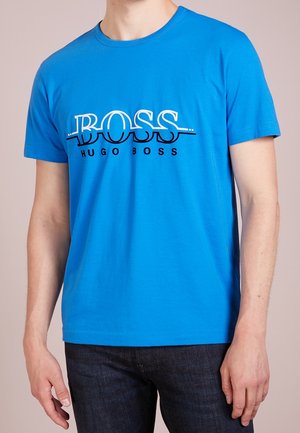 Mand iført en knaldblå kortærmet T-shirt med "BOSS HUGO BOSS" logo i hvidt og sort over brystet, kombineret med mørke jeans.