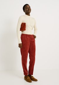 Mujer vestida con un suéter de cuello alto de canalé en color crema, pantalones de pana rojos, mocasines marrones y llevando un pequeño bolso de color óxido bajo el brazo.