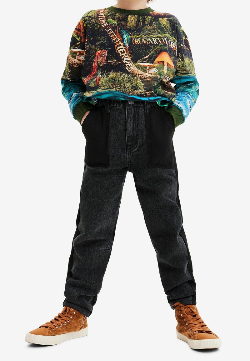 Bluza graficzna z nadrukiem przyrodniczym, w odcieniach zieleni, niebieskiego i pomarańczowego. Czarne spodnie dżinsowe z detalami na bokach i brązowe tenisówki.