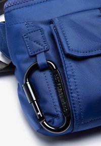 Blå nylon taske med en sort karabin, strukturerede syningsdetaljer og en frontlomme med klap. Glat overflade med funktionelt design.