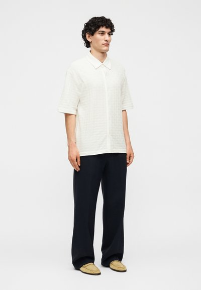 Άνδρας με σγουρά σκούρα μαλλιά, που φοράει λευκό υφασμάτινο κοντομάνικο πουκάμισο, μαύρο παντελόνι με φαρδιά μπάτζια και μπεζ μοκασίνια, στέκεται μπροστά σε απλό φόντο.