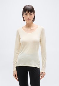 Γυναίκα με κοντά σκούρα μαλλιά που φοράει κρεμ μπλούζα με μακριά μανίκια και V λαιμόκοψη και μαύρο παντελόνι, στέκεται μπροστά σε απλό ανοιχτόχρωμο φόντο.