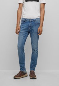 Vaqueros de denim azul claro con un corte Slim, cinco bolsillos y costuras en contraste. Llevados con una camiseta blanca y zapatos de senderismo marrones.