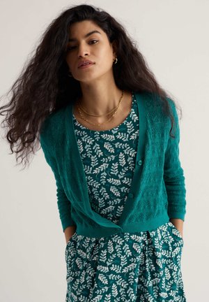 Frau mit langen lockigen Haaren, die goldene Creolen, geschichtete goldene Halsketten, eine tealfarbene Strickjacke und ein passendes, mit Blattmuster bedrucktes Kleid trägt, mit den Händen in den Taschen.