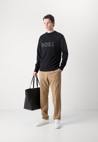 Zwart sweatshirt met 'BOSS' logo in grijs, beige broek, witte sneakers en een zwarte leren tote bag. Eenvoudig, modern ontwerp.