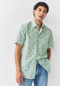 Joven con cabello rizado que lleva una camisa de botones de manga corta color verde menta con pequeños estampados de fresas rojas y jeans azules.