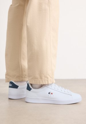 Personne vêtue d’un pantalon beige et de baskets blanches Lacoste avec un détail bleu marine au talon et un petit logo crocodile sur le côté, debout sur un sol clair.