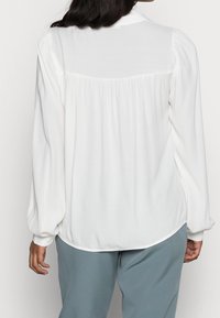 Blusa bianca con maniche lunghe a sbuffo, texture morbida, dettagli plissettati nella parte posteriore e orlo arrotondato, abbinata a pantaloni azzurri chiari.