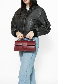 Borsa a mano in pelle rossa con una texture liscia, forma rettangolare e chiusura a pattina. Indossata a mano, insieme a una giacca bomber grigia e jeans chiari.