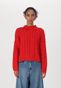 Rød cable-knit genser med høy krage og lange ermer, teksturert mønster, kort lengde, i kombinasjon med lyseblå løstsittende jeans.