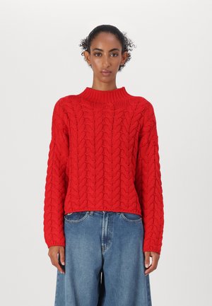 Rød cable-knit genser med høy krage og lange ermer, teksturert mønster, kort lengde, i kombinasjon med lyseblå løstsittende jeans.