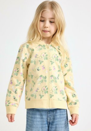 Ung pige med langt blondt hår iført en gul lynlåstrøje med grønne og pink blomster- og bi-mønstre, kombineret med blå jeans.