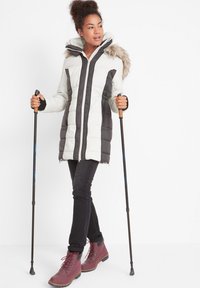 Cappotto imbottito di lunghezza al ginocchio in bianco e grigio scuro con cappuccio in pelliccia sintetica. Il modello tiene delle racchette da trekking, indossando jeans neri e stivali bordeaux.