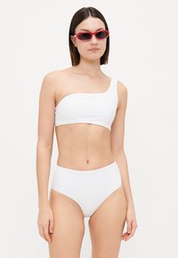 White bikini set featuring a one-shoulder top and high-waisted bottoms. Smooth texture with minimal detailing, paired with pink sunglasses.