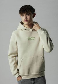 Krem dukserica od mekanog materijala, s prednjim džepom i podesivom kapuljačom. Ispisani natpis "Shady Pines Golf Club" u zelenoj boji.