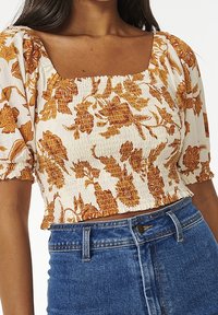 Femme portant un chemisier blanc court avec des imprimés floraux bruns et des manches bouffantes, associé à un jean en denim bleu taille haute.