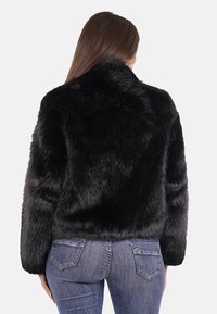 Giacca in pelliccia sintetica nera con colletto alto, superficie testurizzata, maniche lunghe e vestibilità corta, abbinata a jeans blu.
