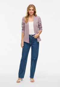 Blazer lila con mangas largas, diseño de frente abierto, usado sobre una blusa blanca y jeans de tiro alto azules. La modelo se encuentra en un fondo neutro.
