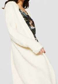 Luddig vit cardigan med en lång, öppen design och vida ärmar, bärs över en blommönstrad topp. Mjuk textur och avslappnad passform.