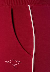 Rote Sporthosen aus weichem Material verfügen über eine seitliche Tasche, die mit weißem Piping eingefasst ist, und ein gesticktes Känguru-Logo in der Nähe der Tasche.