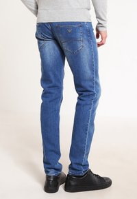 Blue denim jeans with a slim fit, featuring back pockets and subtle stitching details. Paired with black sneakers.