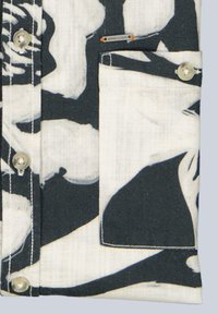 Black and white patterned shirt pocket with beige buttons and a small metal tag above the pocket on light fabric.