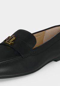 Μαύρο δερμάτινο loafer με χρυσό μεταλλικό έμβλημα στο λουράκι και μπεζ εσωτερική επένδυση σε απλό φόντο.