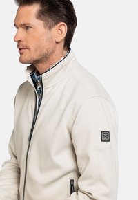 Beige zip-up jack met een gestructureerde stof. Heeft een hoge kraag, zijzakken en een logopatch op de linkermouw.