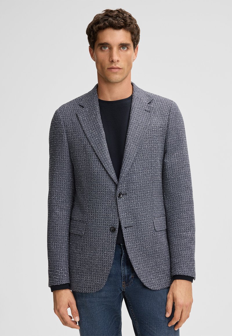 Strukturierter navyblauer Blazer mit hellen Flecken, Einzelknopfverschluss, zwei vorderen Taschen, Reverskragen und figurbetontem Schnitt. Getragen über einem schwarzen Shirt.