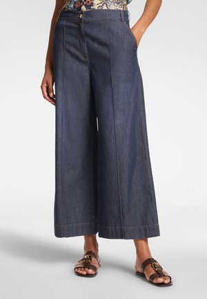 Pantalones de mezclilla de pierna ancha en azul oscuro, con bolsillos frontales, cierre de botón y costuras planas. Combinados con sandalias marrones.