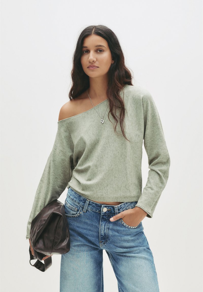 Olivgrünes, schulterfreies Oberteil aus weichem Stoff, kombiniert mit hellblauen Jeans. Dunkelbraune Tasche unter dem Arm gehalten.